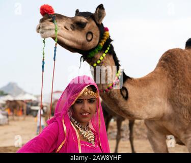 Junge Zigeunerfrau in farbenfroher Kleidung und brillantem Schmuck steht neben dekoriertem Kamel zum Verkauf in Pushkar, Rajasthan, Indien. Stockfoto