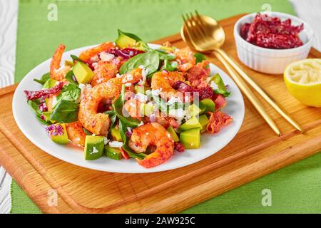 Garnelen-Avocado-Spinatsalat mit sonnengetrockneten Tomaten, zerbröckelten Käse, roter Zwiebel auf einem Teller auf einem Holzbrett, horizontaler Blick von oben Stockfoto