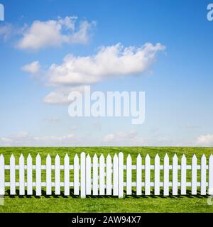 Ein weißer Dickdarmzaun und ein geschlossenes Tor, das im Sommer zu einem schönen grünen Feld und blauem Himmel führt Stockfoto