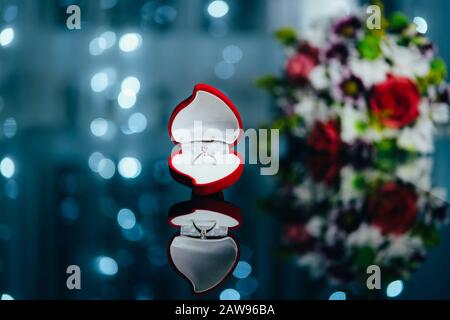 Rote, herzförmige Samtbox mit eingehöhltem Rautenring im Hintergrund mit Bokeh-Lichtern und Blumen. Valentinshintergrund. Geschenke für Frau am Valentinstag. Liebe, Angebotskonzept. Stockfoto