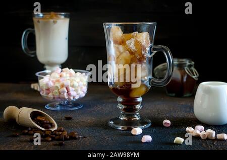 Sommerlicher Eiskaffee in hohen Gläsern mit Eiswürfeln, Milch oder Creamer und dekorierten Marshmallows, braunem Hintergrund und selektivem Fokus Stockfoto
