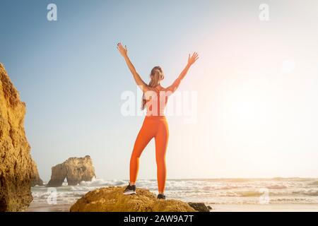 Rückansicht einer fit-Frau mit orangefarbener Sportbekleidung, die auf einem Felsen am Meer steht und mit ihren Armen in Feier, Freiheit angehoben wird. Stockfoto