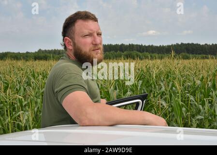 Bauer in der Nähe des Maisfeldes im Sommer. Ein Mann auf dem Hintergrund eines Feldes mit einem Maiswagen weiß. Ein Mann mit Bart auf dem Hintergrund eines Maisfeldes in su Stockfoto