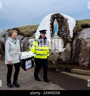 Garda Tom McBride mit Presiding Officer für Gola Island Nancy Sharkey, tragen eine Wahlurne, die die Statue der Jungfrau Maria am Hafen der Insel vor der Küste von Donegal, Irland, passiert. Auf das Festland zurückzukehren, nachdem Islanders bei der irischen Wahl einen Tag vor dem Rest des Landes ihre Stimme abgegeben hatte. Stockfoto