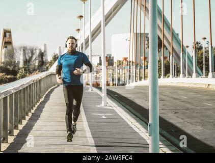 Junger Mann, der auf einer Brücke in einem blauen Hemd läuft und mit einem Paar Kopfhörern Musik hört. Stockfoto