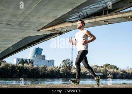 Junger Mann, der in einem weißen Hemd über eine Brücke läuft. Er hört Musik, und er hat ein paar Helme auf. Stockfoto