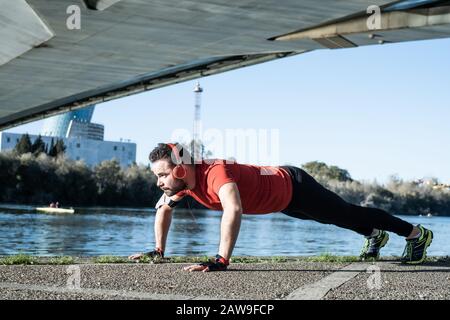 Junger Mann, der mit einem roten T-Shirt und einem Musikkopfhörer in urbaner Umgebung Push-ups macht. Gesundes Wohnkonzept Stockfoto