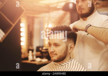 Brutaler Barber, der modisches Styling von bärtigen Männern in Cape macht. Im Retro-Stil des Beauty-Salons verschwommener Hintergrund Stockfoto