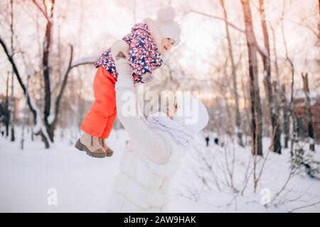 Mutter und Tochter verbringen fröhlich den Wintertag im schneebedeckten Park. Hände an den Schneebällen der Frau. Sie hob ihr Baby in die Luft, Flugsimulatio Stockfoto