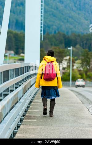 Einheimische Alaska-Frauen tragen eine leuchtend gelbe Regenjacke und einen orangefarbenen Rucksack, der über die O'Connell Bridge in Sitka, Alaska, USA läuft. Stockfoto