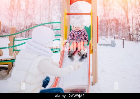 Mama fängt ihre Tochter am Boden der eisernen Rutsche auf dem Spielplatz im Morgengrauen des Winterparks Stockfoto