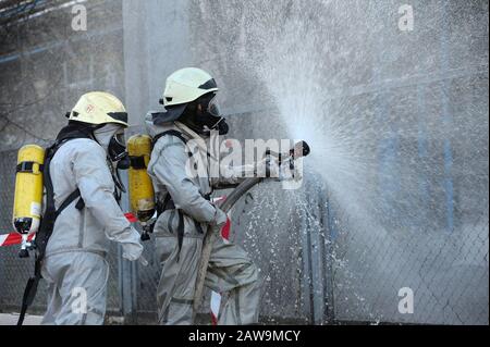 Rettungskräfte in schützenden Gummianzügen bewässern Pflanzengebiet mit Spritze. Schulung des Rettungsteams in der Dekontamination Stockfoto