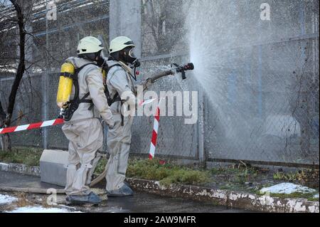 Rettungskräfte in schützenden Gummianzügen bewässern Pflanzengebiet mit Spritze. Schulung des Rettungsteams in der Dekontamination Stockfoto