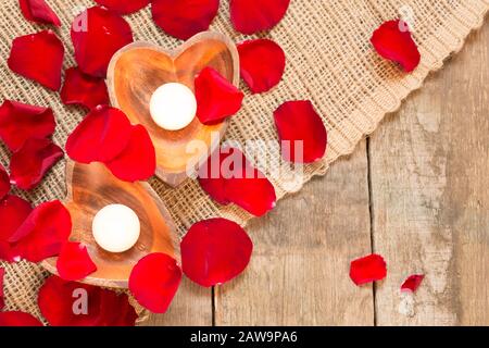 Erleuchtete Kerzen in herzförmigen Kerzenhaltern mit roten Rosenblättern auf rustikalem Holzhintergrund. Hintergrund des St Valentines. Romantisches Urlaubskonzept. Draufsicht. Horizontal Stockfoto