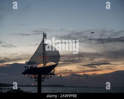 Ein Schild mit Metall-Jacht aus Cobes Gurnard in der Profilsilhouette gegen den Himmel bei Sonnenuntergang über dem Solent mit nach rechts fliegendem Vogel Stockfoto