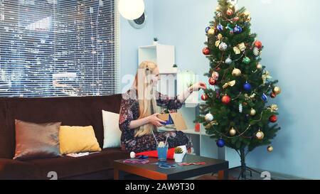 Hübsches Mädchen hängt Spielzeug am Weihnachtsbaum mit Wolkenkratzern auf dem Hintergrund und sitzt auf einem Sofa mit Kopfkissen zu Hause. Sie nimmt die Spielzeuge aus der Schachtel. Stockfoto