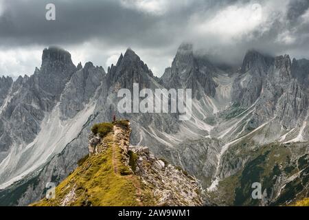 Mann mit roter Jacke, der auf einem Grad steht, hinter ihm Berggipfel und Spitze Felsen, dramatische Wolken, Cimon der Croda Liscia und Cadini Gruppe Stockfoto