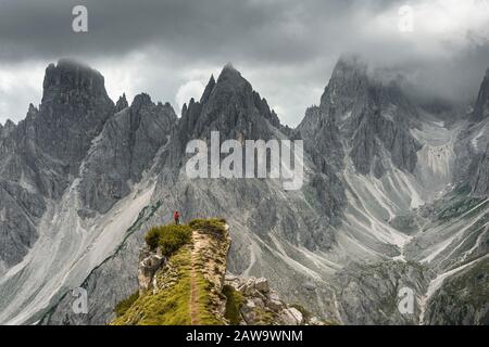 Mann mit roter Jacke, der auf einem Grad steht, hinter ihm Berggipfel und Spitze Felsen, dramatische Wolken, Cimon der Croda Liscia und Cadini Gruppe Stockfoto