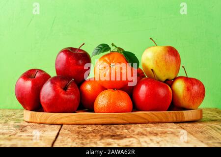 Rote reife Äpfel und Mandarinen mit grünen Blättern liegen auf einem Holztablett auf einem alten Holztisch auf hellgrünem Hintergrund, Bild mit Kopierraum. Stockfoto