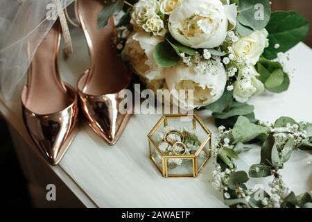 Hochzeitszubehör auf dem Schminktisch: Ein brautstrauß aus weißen Ponys, Hochzeitsringe in einer Glaskiste, Goldschuhe. Vorbereitung auf die Hochzeit am Morgen. Stockfoto