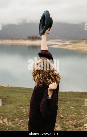 Schöne Frau im violetten Pullover, die ihren Hut hält und eine Reise auf dem See genießt. Weibliche Touristen, die den See erkunden. Sie lächelt und fängt ihren Hut. Stockfoto