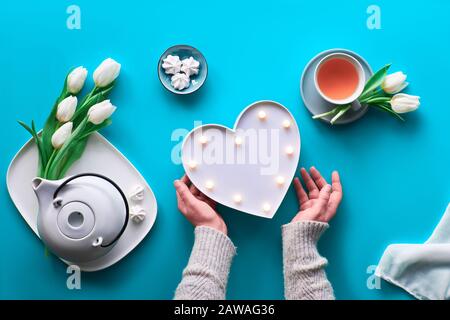 Frühlingsfeier flach lag. Weibliche Hände zeigen Herzformzeichen. Teetasse, Teekanne, Süßigkeiten und weiße Tulpen auf blauem Tisch. Muttertag, international w Stockfoto