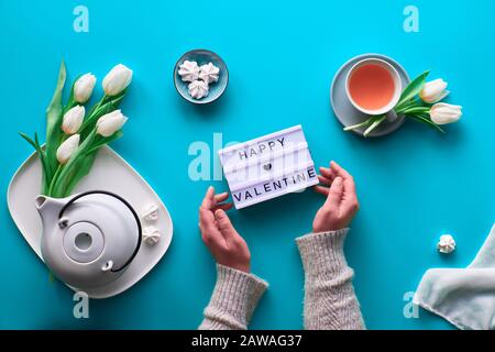 Flaches Federmuster. Weibliche Hände halten Leuchtkasten mit Text "Happy Valentine". Teetasse, Topf, Süßigkeiten und weiße Tulpen auf Blau. Valentinstag Cele Stockfoto