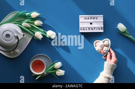 Frohe Festtage zum Valentinstag Wohnung lag. Weibliche Hände zeigen Herzformzeichen. Teetasse, Teekanne, Süßigkeiten und weiße Tulpen auf Geschichte, Hintergrund im Trend Stockfoto