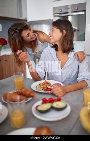 Zwei Frauen lächeln, umarmen, essen, reden in der Küche, unterhalten. Freizeit, Wohnen, modern, Lifestyle, Konzept Stockfoto