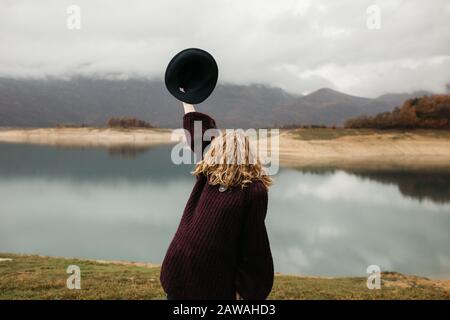 Fröhliche schöne Frau im violetten Pullover, die ihren Hut hält und eine Reise auf dem See genießt. Sie lächelt und fängt ihren Hut. Stockfoto