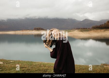 Fröhliche schöne Frau im violetten Pullover, die ihren Hut hält und eine Reise auf dem See genießt. Sie lächelt und fängt ihren Hut. Stockfoto