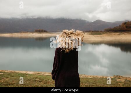 Glückliches Mädchen mit lockig blonden Haaren tanzt allein auf einem See, ihr Haar flieht wegen des Windstroms, frei als Vogel. Foto von Mädchen mit lockigem Haar ST Stockfoto