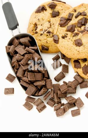 Dunkle Schokoladensplitter in einer silberfarbenen Küchenschaufel. Im Hintergrund befinden sich gestapelte Schokoladen-Chip-Plätzchen. Stockfoto