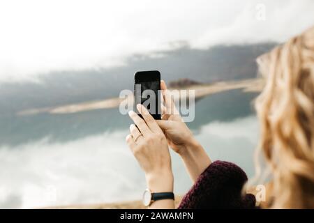 Nahaufnahme der weiblichen Hände, die das Handy halten und an einem bewölkten Tag Fotos vom See machen. Sie textet auf Smartphone und nimmt selfie mit. Stockfoto