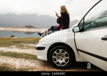 Schöne Frau mit lockigem Haar, sitzt auf dem Auto, hält Handy und fotografiert an einem trüben Tag den See. Sie textet auf dem Smartphone Stockfoto