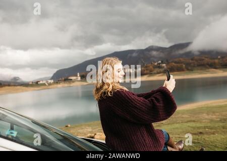 Schöne Frau mit lockigem Haar, sitzt auf dem Auto, hält Handy und fotografiert an einem trüben Tag den See. Sie textet auf Smartphone und Stockfoto