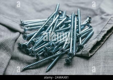 Eine Handvoll metallischer selbstschneiderschrauben liegt vor dem Hintergrund eines alten Denim-Stoffes. Makroaufnahme mit selektivem Fokus und flachem Freiheitsgrad Stockfoto