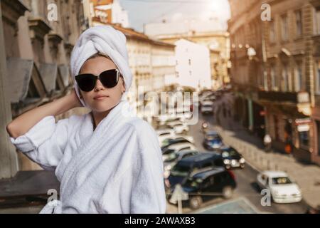 Mädchen in einem weißen bademantel und einem Handtuch auf dem Kopf in Sonnenbrille steht auf dem Balkon vor dem Hintergrund der Stadt Stockfoto