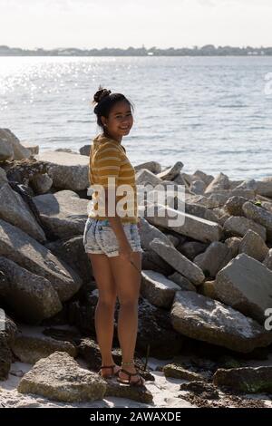 Eine schöne junge Frau in einem gelb gestreiften Top steht in der Nähe eines Schutterhaufen entlang Indian River Lagoon auf Hutchinson Island in Stuart, Florida, USA. Stockfoto