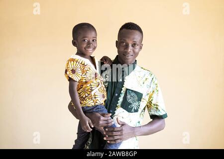 Afrikanischer Vater posiert stolz mit seinem Sohn vor gelblichem Hintergrund lächelnd und lacht mit Kind Stockfoto