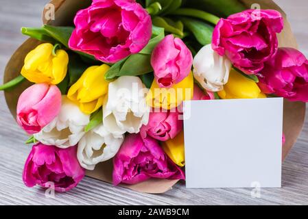 Hallo Frühlings-Zeit-Konzept. Nahaufnahme von wunderschönem Blumenstrauß aus buntfarbenem, pink-gelb-weißen Blumen in Papierverpackung mit Postkarte auf Grau liegend Stockfoto