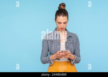 Porträt einer ernsthaften, fokussierten Frau mit Haarbun im modischen Outfit, das auf dem Smartphone tippt, im sozialen Netzwerk textet, im Internet stöbert, mit mobilem Phon Stockfoto
