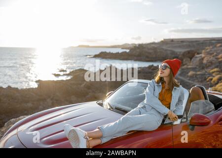 Junge stilvolle Frau genießen schöne Landschaften, liegen auf der Autohaube während der Reise auf der felsigen Küste bei einem Sonnenuntergang. Unbeschwertes Lifestyle- und Autoreisekonzept Stockfoto