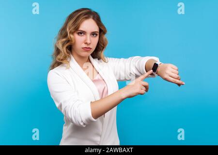 Mal höher! Porträt einer ernsthaften Frau mit gewelltem Haar in weißer Jacke, das auf die Uhr auf der Hand zeigt und enttäuschend aussieht, unzufrieden mit der Verspätung. Stockfoto