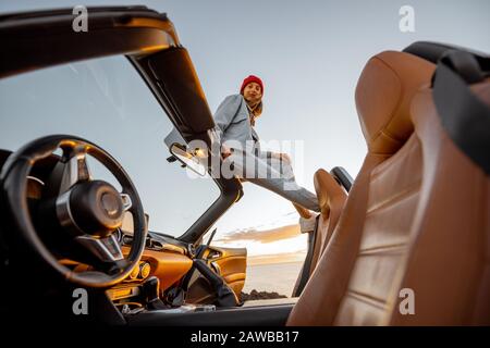 Frau, die bei Sonnenuntergang auf dem Dach des Autos sitzt und einen schönen Blick auf das Meer genießt. Naturgenuss und unbeschwerte Reisen mit dem Autokonzept Stockfoto