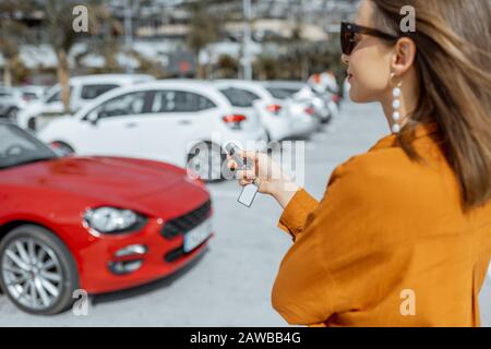 Junge Frau, die Schlüsselanhänger eines neuen gekauften oder gepachteten roten Autos auf dem Parkplatz im Freien hält Stockfoto