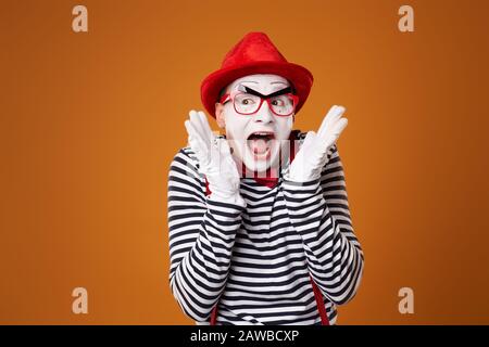 Überraschter junger MIME-Mann in rotem Hut und Weste auf leerem orangefarbenem Hintergrund Stockfoto