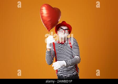 Hocken von MIME Clown in rotem Hut und Weste hält Herzball in seinen Händen auf leerem orangefarbenem Hintergrund Stockfoto