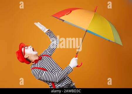 Trauriger MIME-Mann mit mehrfarbigem Regenschirm in der Hand, isoliert auf leerem orangefarbenem Hintergrund im Studio Stockfoto