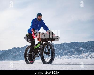 Ein männlicher Reisender fährt mit einem Fatbike auf einem gefrorenen Bergsee. Reisen im Winter. Extreme Expedition. Stockfoto